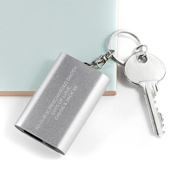 A personalised silver aluminium emergency power bank keyring engraved with 'Drive safe. I need you here with me. Love Sarah x', lying on a dark textured surface next to a smartphone.