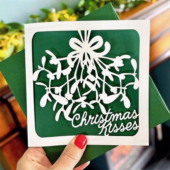 Hand holding a white mistletoe papercut Christmas card with a forest green paper insert and matching green envelope