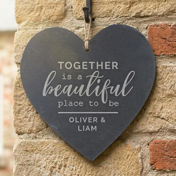 A dark grey heart-shaped slate plaque engraved with 'Together is a beautiful place to be' hanging from a black metal hook on a rustic stone and brick wall.
