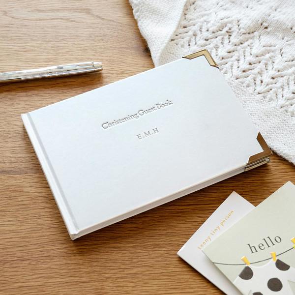 Personalised Christening Guest Book in white with silver corners, silver-foil engraved with initials E.M.H on a wooden surface.