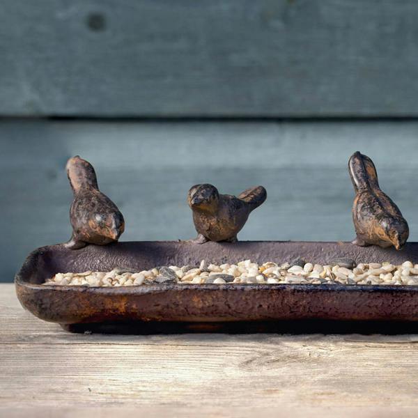 A rustic, heavy-duty cast iron feeding tray with a textured, weathered finish resting on a light wooden surface. Three decorative metal bird figurines are perched on the edges of the tray, which is filled with a mixture of bird seeds and grains.