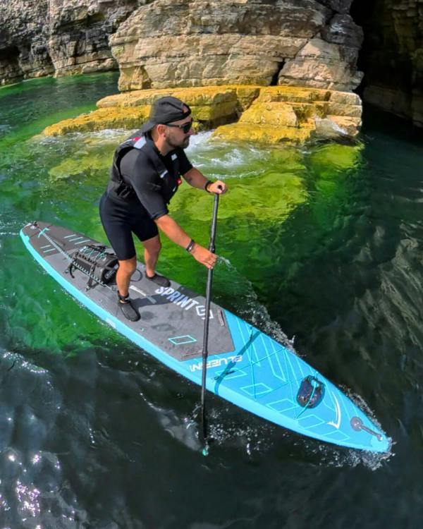 Paddleboarder in black wetsuit navigating clear green waters near rocky cliffs.