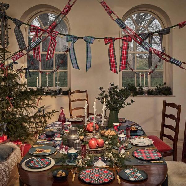 A festive dining room decorated with a tartan bow garland draped across arched windows above a dining table set with plaid plates.