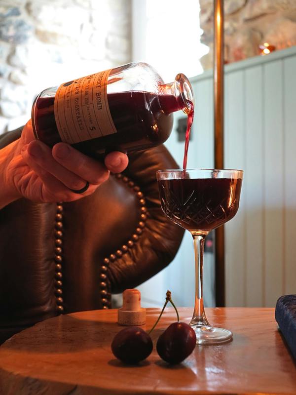 A hand holds a glass bottle 'Cherry Bomb' by 'Prohibition Cocktails', pouring a dark red liquid into a coupe glass on a wooden table. Two dark cherries and a cork are visible on the table, with a brown leather chair and softly blurred indoor background.