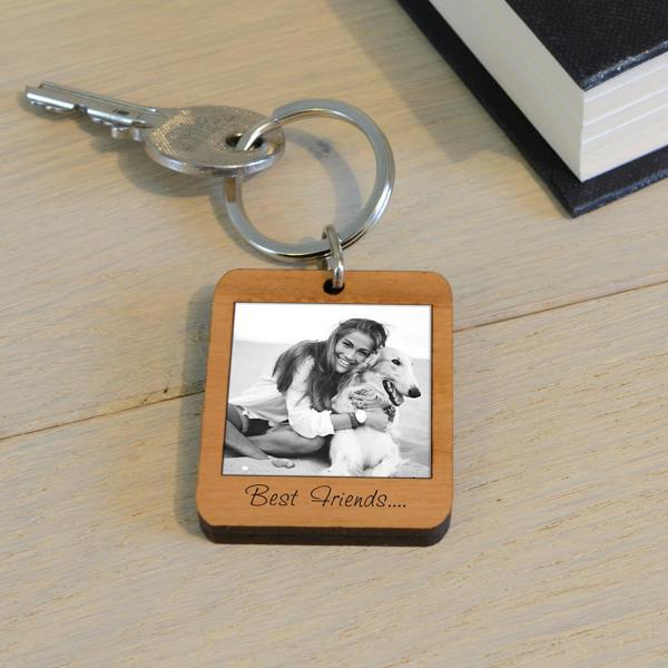 A rectangular solid cherry wood keyring cut into a classic Polaroid style frame, featuring a printed black and white photo of a smiling girl hugging her white dog. Engraved text below the photo reads "Best Friends....". The keyring sits on a light wooden desk next to a silver key and a black book.