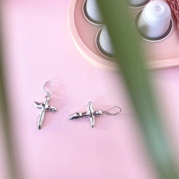 Silver cross earrings photographed on pink surface with palm leaf.