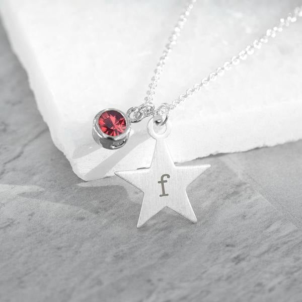 A close-up shot of a delicate silver chain necklace resting on a white textured stone, featuring a small brushed-silver star pendant engraved with a lowercase letter 'f' alongside a small round ruby-red crystal charm.