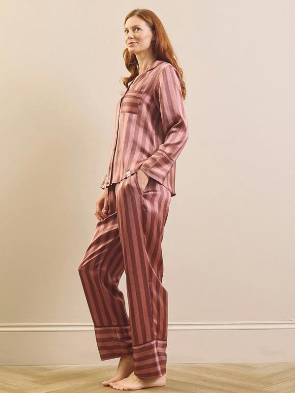 A woman modelling a matching two-piece pyjama set with vertical stripes in dusty rose and dark brown, standing barefoot against a neutral beige wall.