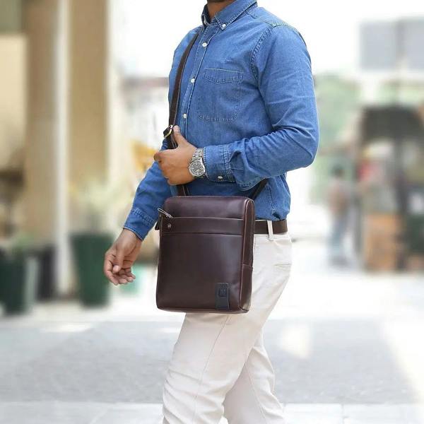 Man wearing a blue shirt and white pants with a brown leather messenger  bag on a blurred background