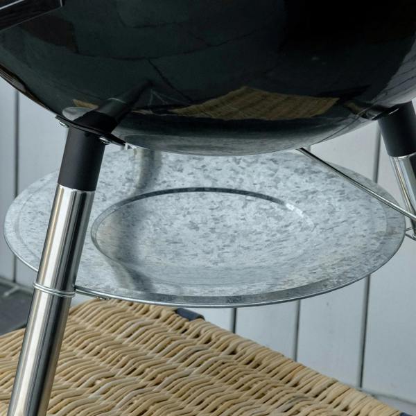 Metal ash tray at the bottom of a black kettle charcoal grill with charcoal inside.