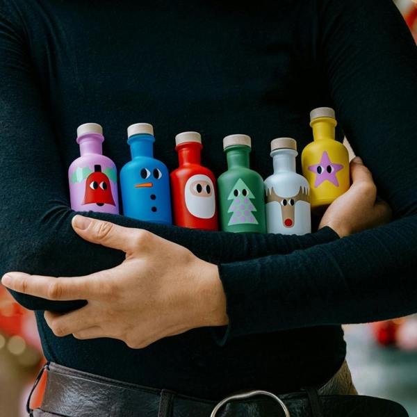 A close-up of a person cradling six small, colourful Christmas-themed olive oil bottles with wooden stoppers against a soft-focus background of festive lights.