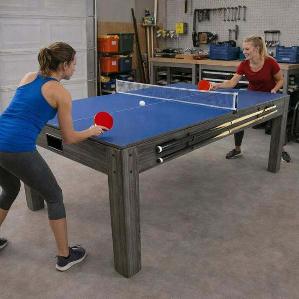7-in-1 games table with a blue surface, sturdy grey wood frame, and two players enjoying a game of table tennis in a garage setting