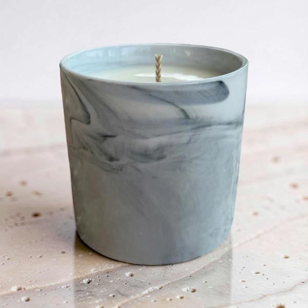 A gray and white marble-patterned glass candle jar with a single wick, set on a light stone surface against a soft neutral background.