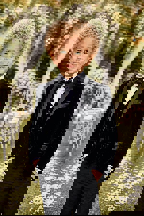 Child in a black tuxedo with a bow tie standing outdoors with floral decorations.