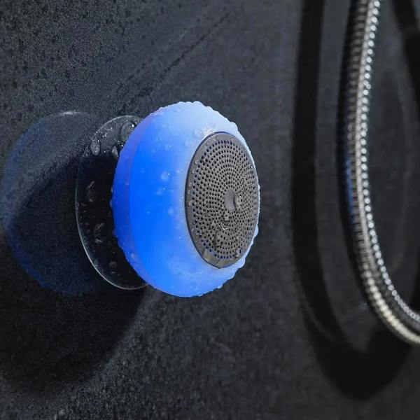 A round, blue waterproof Bluetooth shower speaker attached to a dark, wet shower wall via a suction cup, with the speaker casing glowing blue.