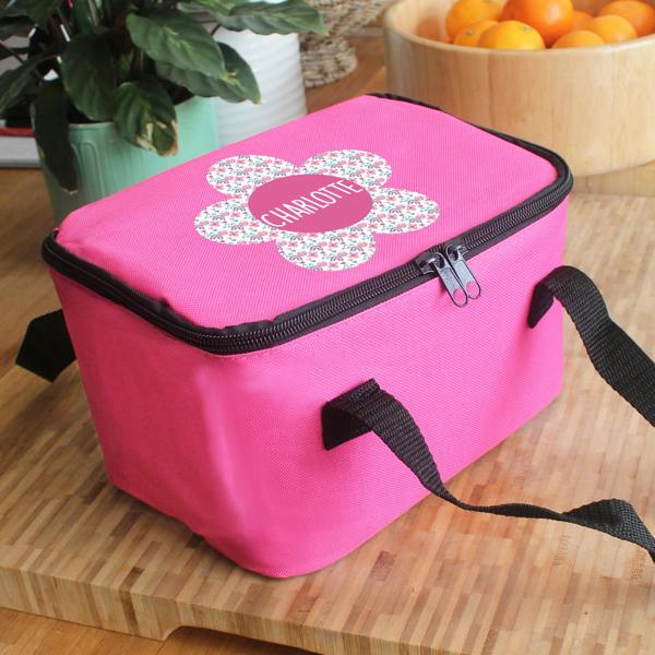 An insulated pink children's lunch bag custom printed with the name Charlotte inside a central patterned flower motif, sitting angled on a wooden surface.