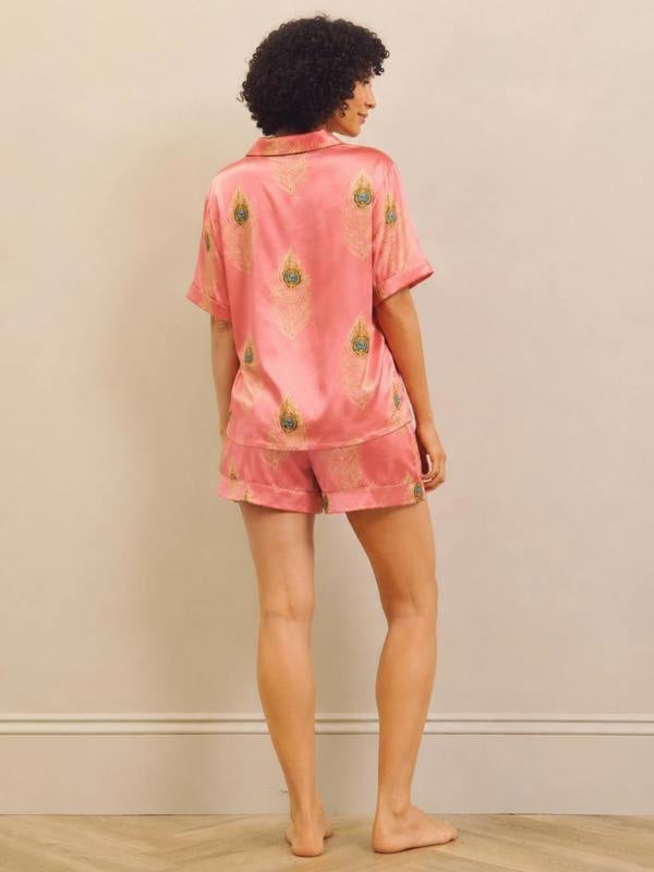 A woman stands barefoot against a plain beige wall, viewed from the back, wearing a pink short-sleeve pyjama top and matching shorts with a gold and green peacock feather pattern.