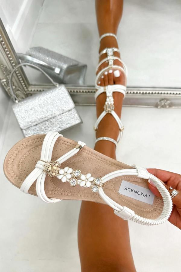 White strappy flat sandals with pearl and crystal embellishments on the toe strap, shown worn on a foot and held in hand against a light tiled background with a mirror reflection.