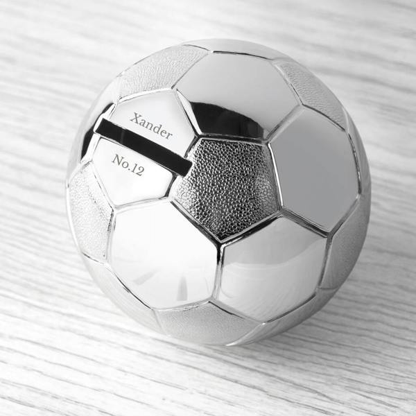 Personalised silver-plated football money box showing engraved custom name text above and below the coin slot on a light wood background.