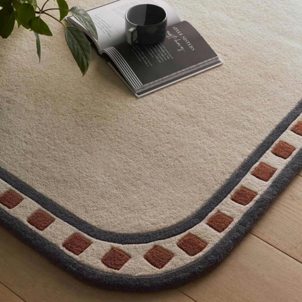 Decorative rug with a geometric border on a wooden floor, featuring a book and cup.