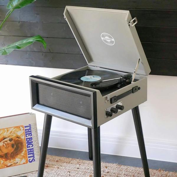 A retro style grey record player on black legs, featuring an open lid with a vinyl record playing, styled in a modern home interior.