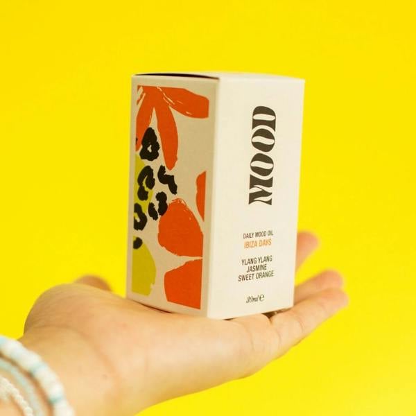 Hand holding the illustrated Ibiza Days Daily Aromatherapy Mood Oil boxed packaging against a bright yellow background by Abode Home & Gifts