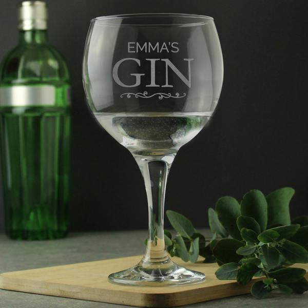 A personalised stemmed gin balloon glass by Abode Home & Gifts, featuring the custom laser-engraved text "EMMA'S GIN" above a decorative scroll, filled with a clear gin drink on a wooden serving board next to a green gin bottle and leafy eucalyptus sprigs.