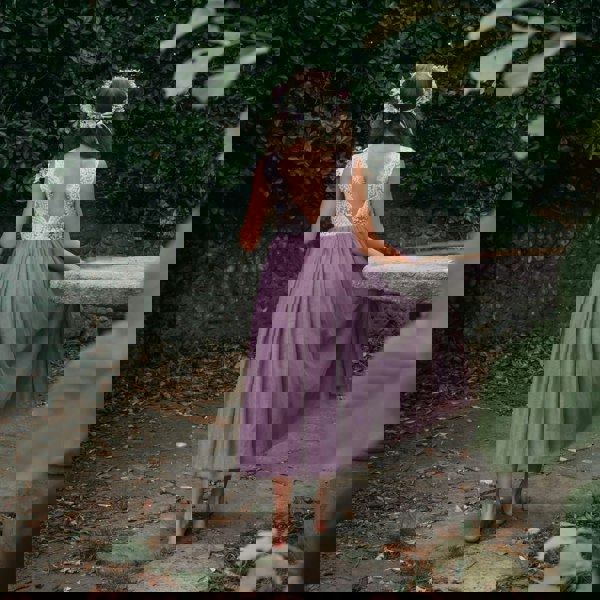 Girl in a purple dress with a floral headband walking away from the camera in a garden setting.