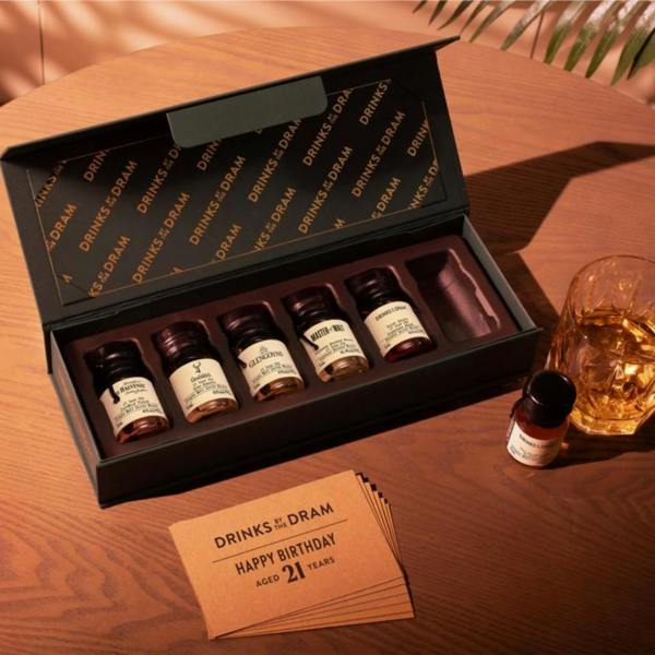 An open premium black gift box from Abode Home & Gifts displaying six wax-sealed 21-year-old whisky drams on a wooden table, next to a crystal tumbler of whisky and a 21st birthday card.