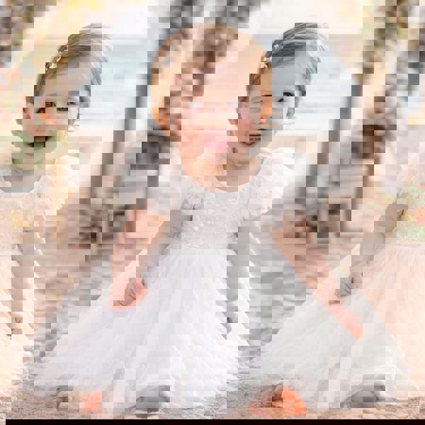 Baby in a white dress sitting outdoors with flowers and a bench in the background