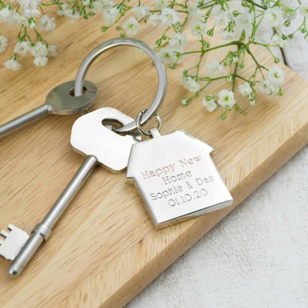 A silver house-shaped keyring engraved with the text "Happy New Home", "Sophie & Dan", and "01.10.20", resting on a light wooden board next to a set of house keys and decorative baby's breath flowers.