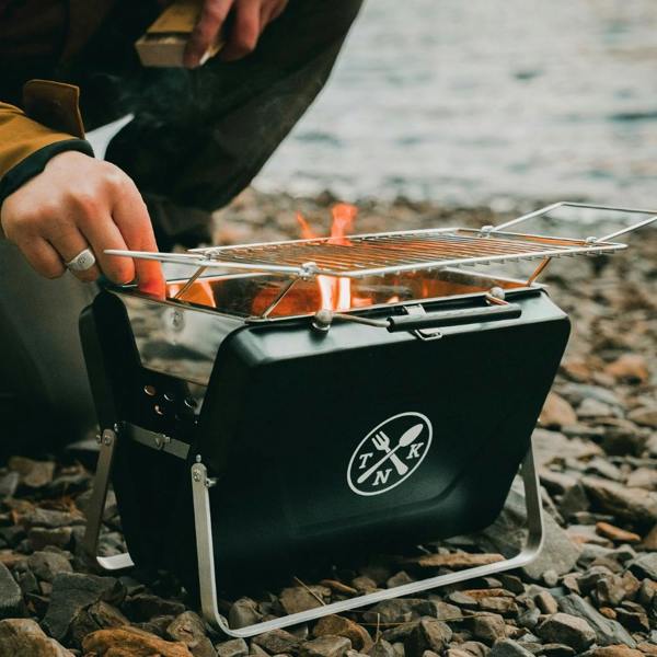 Person setting up the Portable Notebook BBQ with a fire starter on a pebble beach.