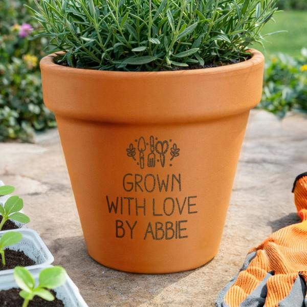 A personalised terracotta plant pot engraved with 'Grown With Love By Abbie' holding a lush green plant on an outdoor stone patio beside garden gloves.