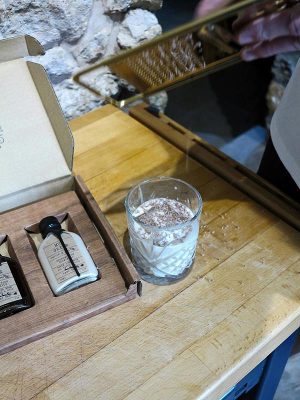 A small clear glass tumbler filled with a white beverage and topped with cocoa powder, set on a wooden table next to an open gift box containing two cream-colored bottles. A hand is holding a gold grater above the glass.