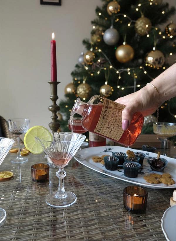 A hand pours a pinkish-red liquid from a bottle into a coupe glass on a decorated holiday table with a Christmas tree in the background.
