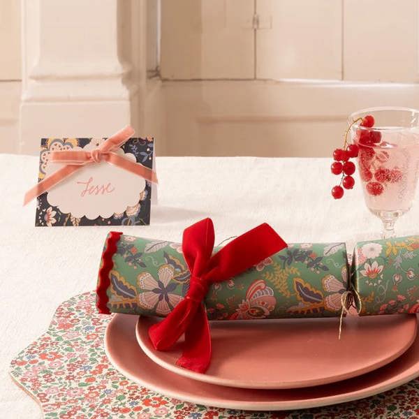 An elegant festive dinner table setting showcasing a luxury floral place card with a pink velvet bow alongside a coordinating pattern cracker, scalloped placemat, and blush pink table linen.
