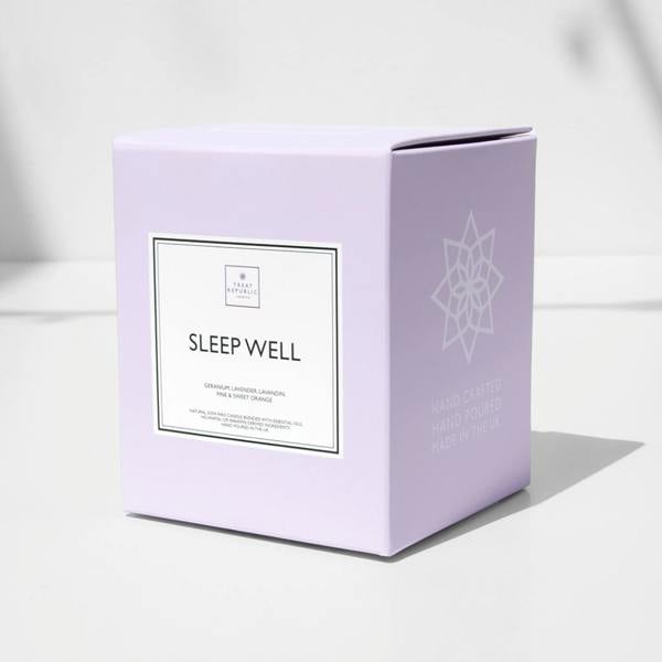 A close-up studio shot of the premium pastel purple retail presentation box for the Sleep Well scented candle. The front of the square box displays a crisp white minimalist label outlining the essential oil ingredients. The side panel features a white geometric star emblem above the printed text 'HAND CRAFTED, HAND POURED, MADE IN THE UK'. The gift box is positioned on a clean white surface with a soft shadow cast to the right.