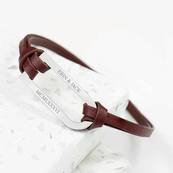 A custom engraved men's brown leather bar bracelet from Abode Home & Gifts reading 'ERIN & JACK' and 'MCMLXXXVI' on a white marble tier.