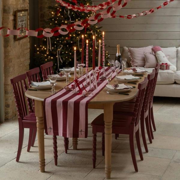 A wooden dining table dressed with a burgundy and pink striped organic cotton table runner, set with red candles and tableware in front of a lit Christmas tree.