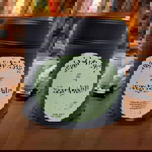 Candle in a tin labeled 'Ralph's Orchard vintage bookshop' on a wooden surface with a blurred background.