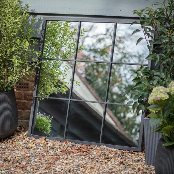 The 90x90cm square black metal window pane mirror leaning on a gravel path against an outdoor wall by Abode Home & Gifts.