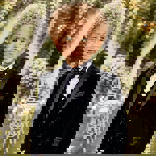 Child in a black tuxedo with a bow tie standing outdoors with floral decorations.