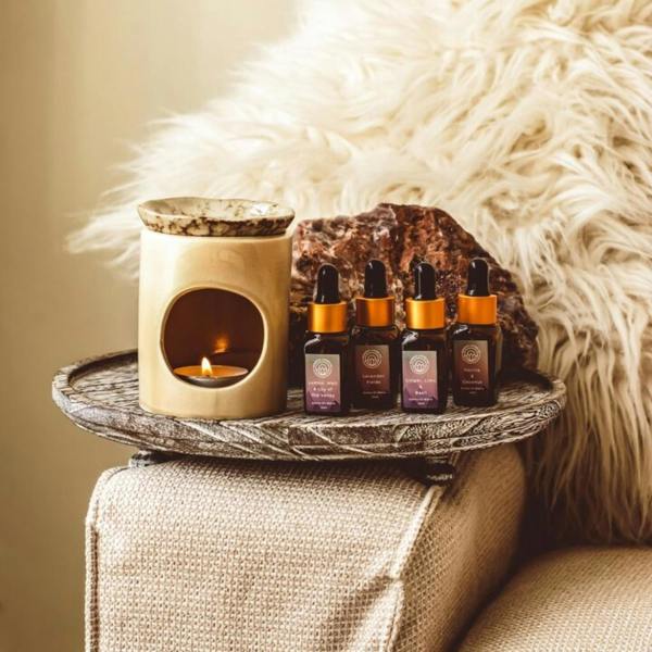 Handmade crackled ceramic oil burner with lit tealight alongside four essential oil dropper bottles on a textured wooden tray next to a cozy armchair by Abode Home & Gifts