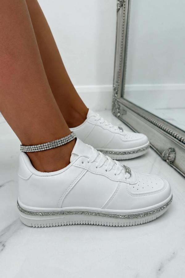 White low-top sneakers with silver rhinestone trim on the soles and laces, shown on a model's feet against a white marble floor and a silver-framed mirror.