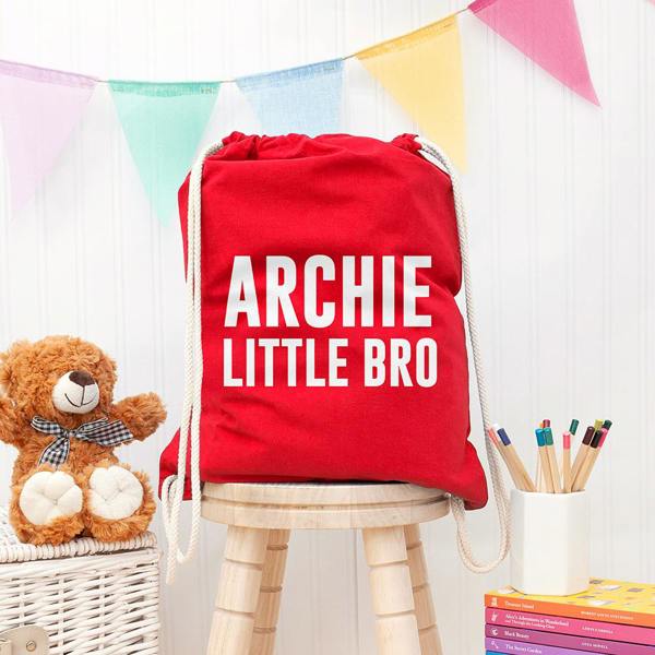 Boy's personalised bright red gym drawstring bag printed in bold white text with ARCHIE LITTLE BRO, resting on a wooden stool.