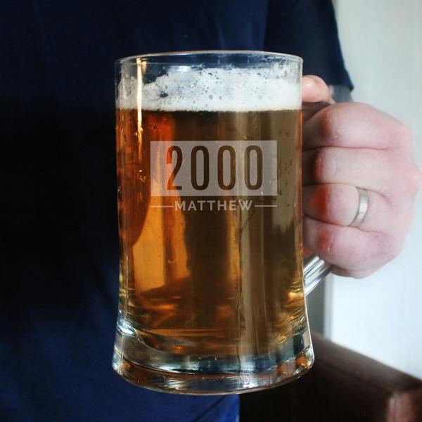 A close-up of a person holding a glass pint tankard filled with amber beer and foam, engraved with 2000 and MATTHEW.