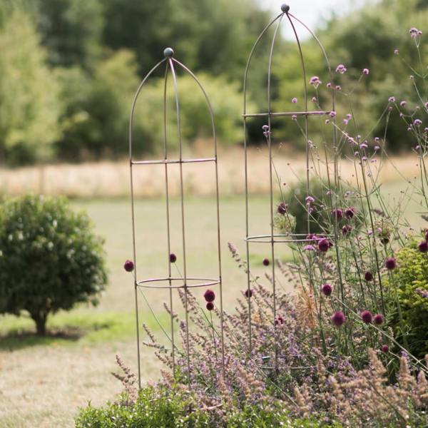 Two tall raw metal garden obelisks of different heights standing in a blooming garden border.