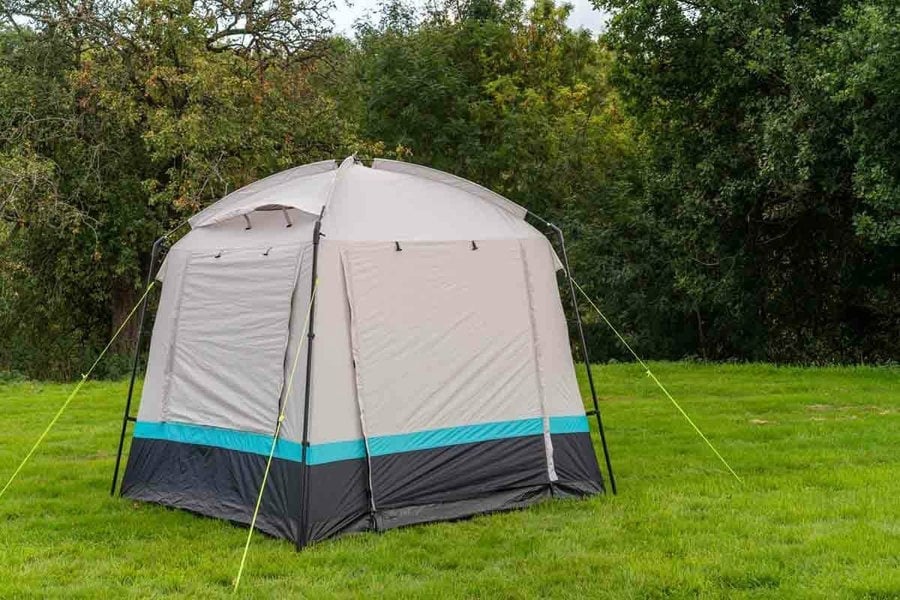 The OLPRO Pod Kitchen/Storage Tent pitched on a campsite with its door closed and a tree line in the background.