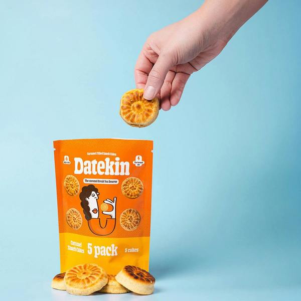 Hand holding a Datekin caramel cake above a pack of caramel cakes with cakes displayed on a light blue background
