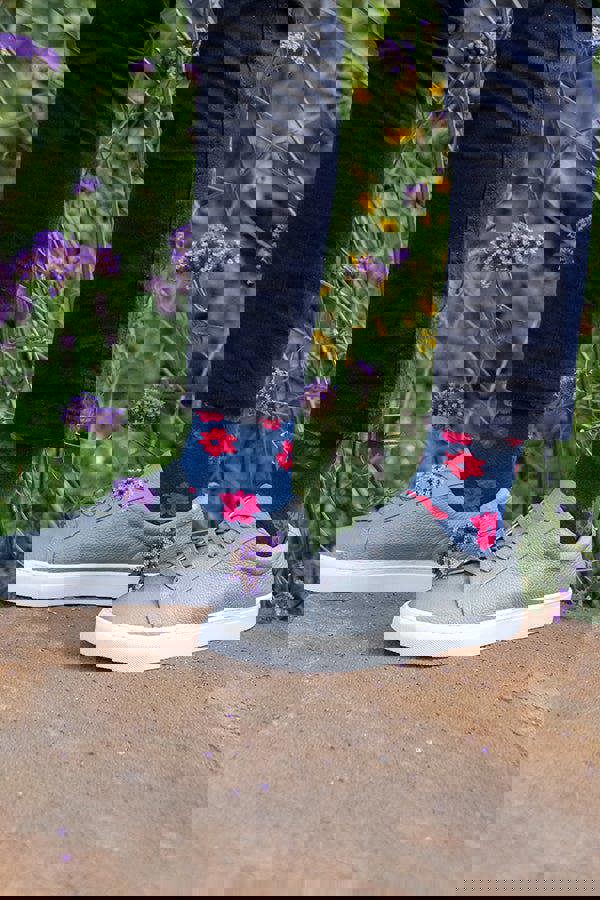 Person wearing blue socks with red floral patterns and gray sneakers standing on a rock with greenery in the background.